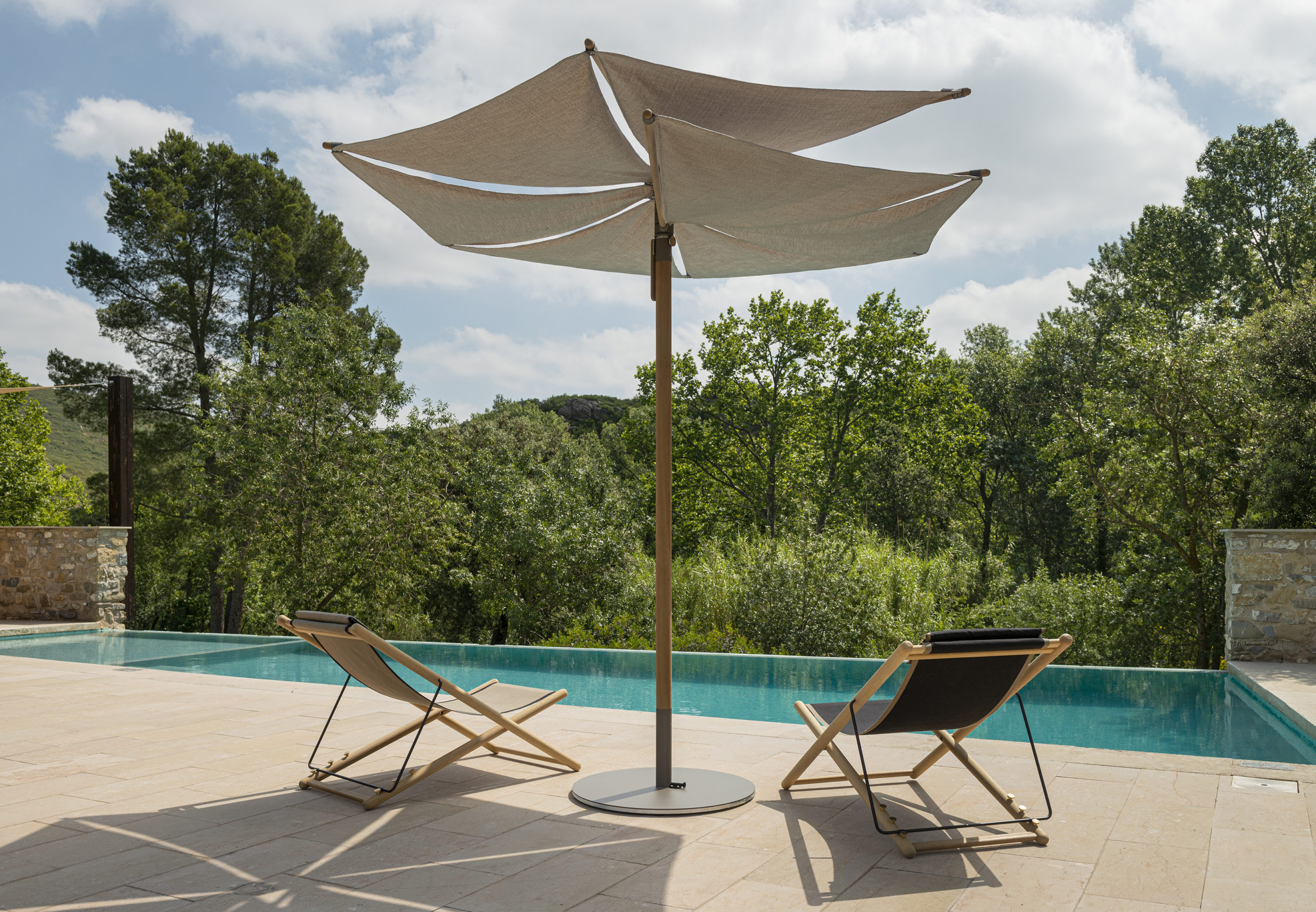 Two wood-framed Om parasols open over a limestone pool terrace in full sun, the teal water of an infinity pool between them and a dry-stone wall with planting beyond.