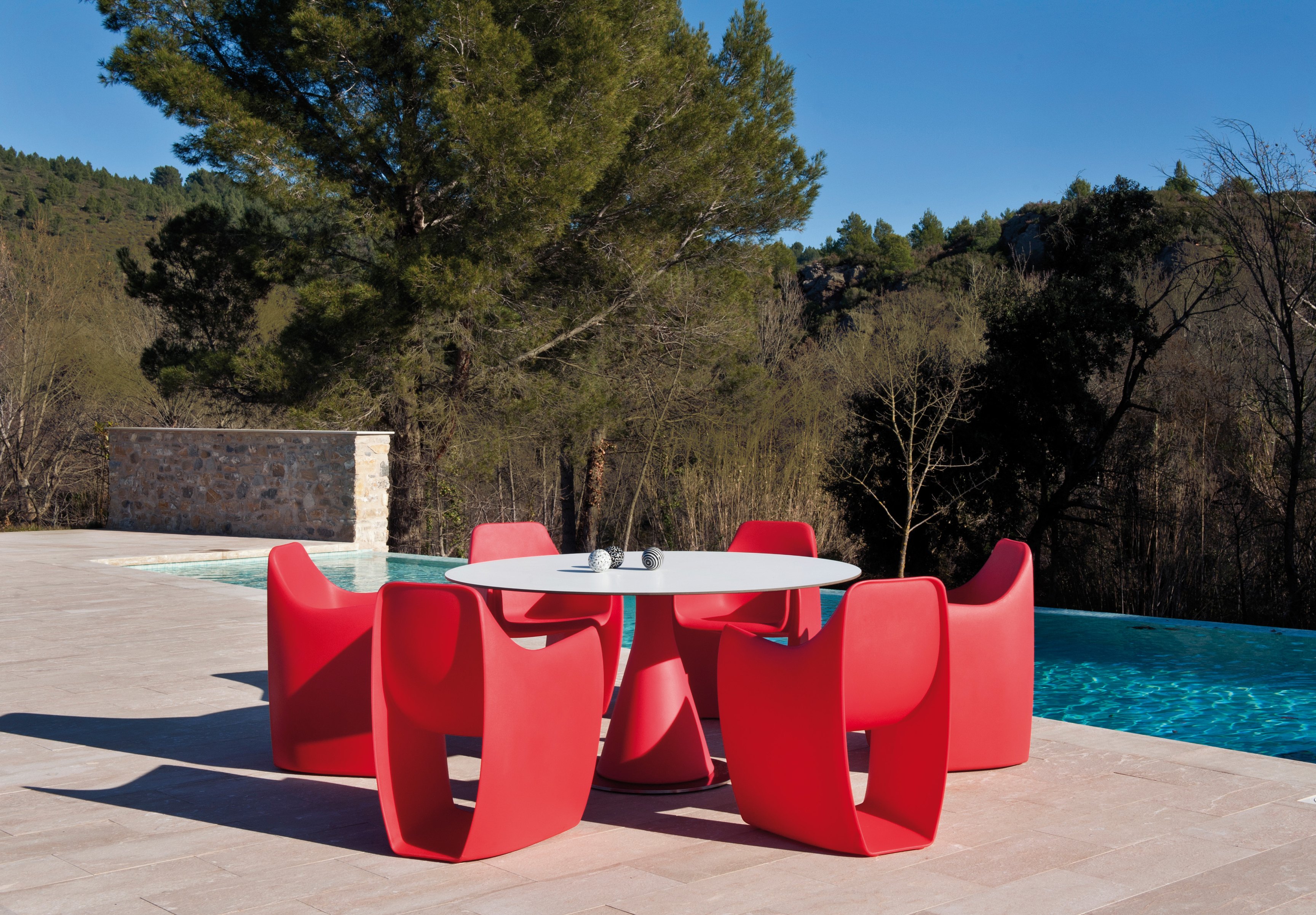 Six red rotomoulded armchairs with cut-away sides around a round white-topped table on a pale stone pool terrace, an infinity pool behind them and a pine wood rising on the hillside beyond.