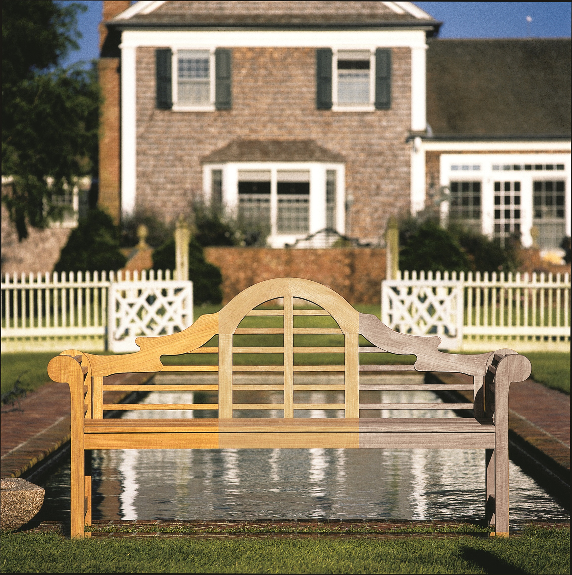 A teak Lutyens bench with an arched back, its left half honey-coloured and its right half weathered to silver-grey, on a lawn before a long reflecting pool, white fretwork gates and a shingled house with dark shutters.