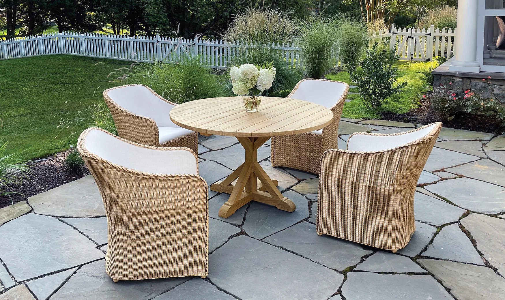 Woven dining armchairs with white cushions round a teak table on a flagstone terrace, white hydrangeas on the table, ornamental grasses and a white picket fence behind.