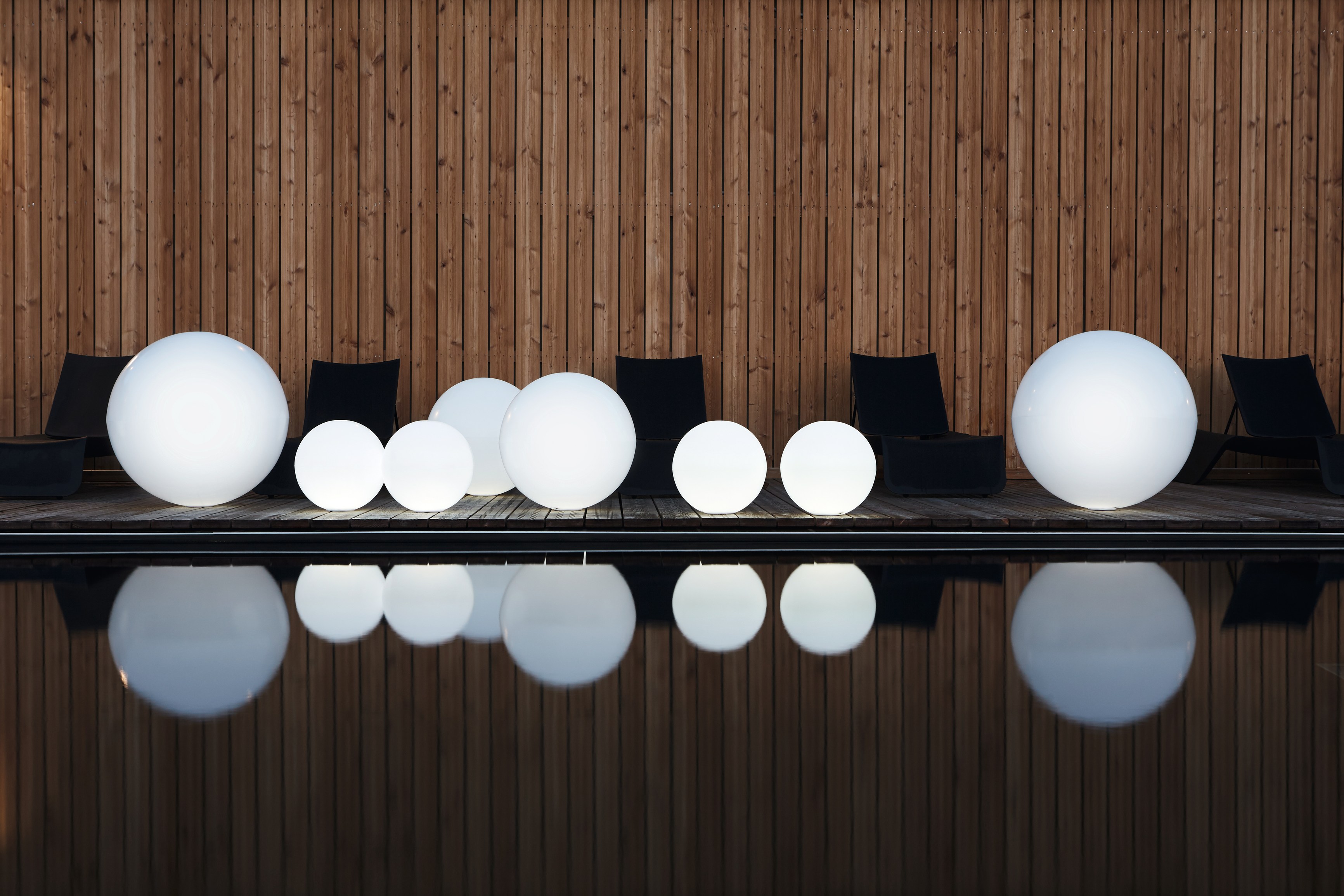 Illuminated white CRYSTAL sphere lights in four sizes ranged along a timber pool deck, black sunloungers set against a vertical-slat timber wall behind them, each globe mirrored in the still dark water at dusk.
