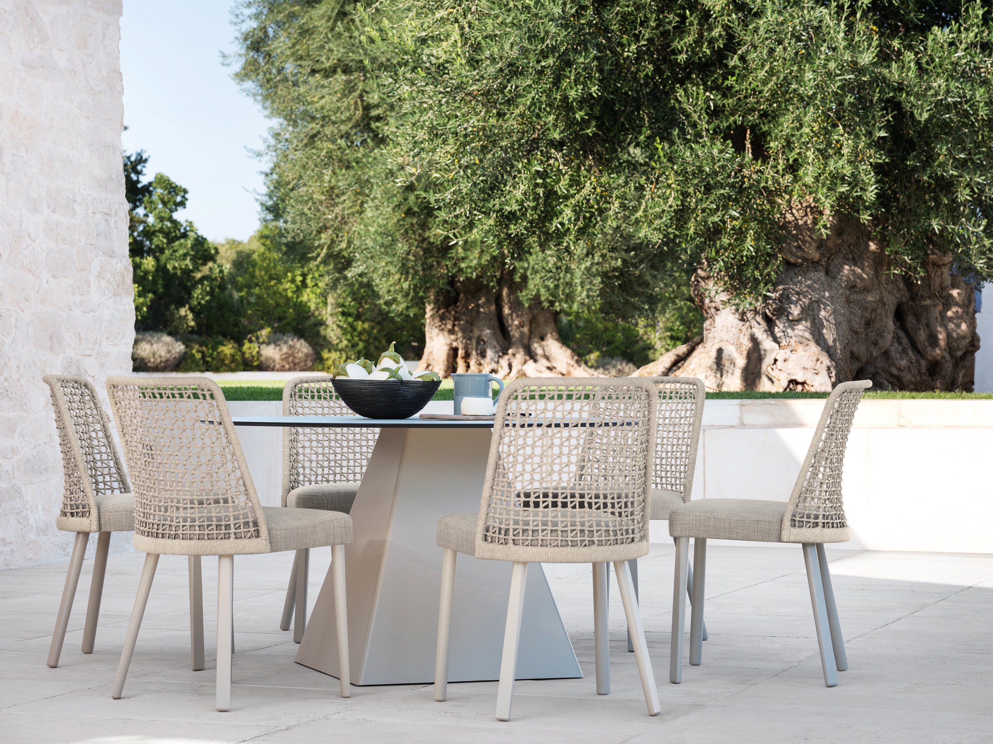 Six rope-woven dining chairs around a round table on a pale stone terrace, two ancient olive trees behind.