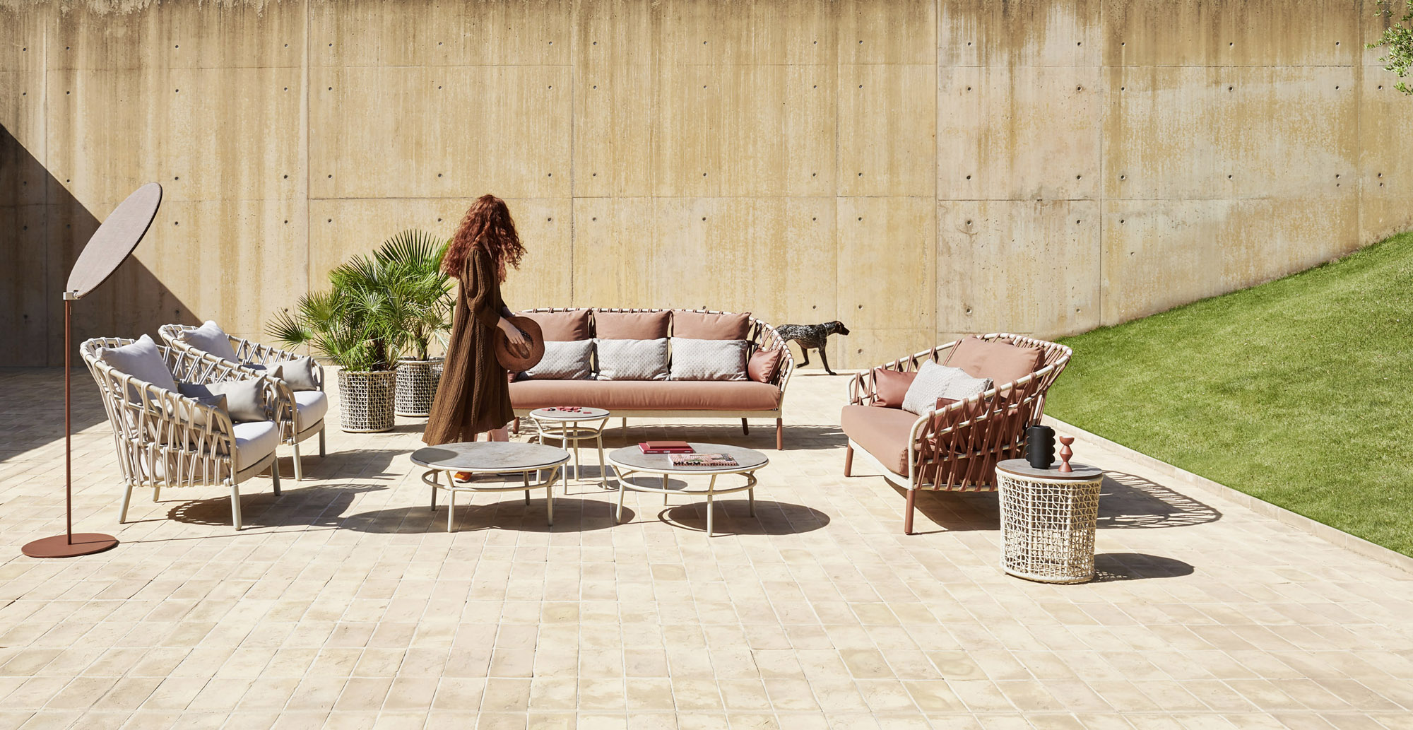 A dusty-terracotta cross-arm sofa and lounge chairs under a bronze-framed cantilever shade on a sunlit terrace, a board-formed concrete wall and potted palms behind, a woman and a dog beside them.