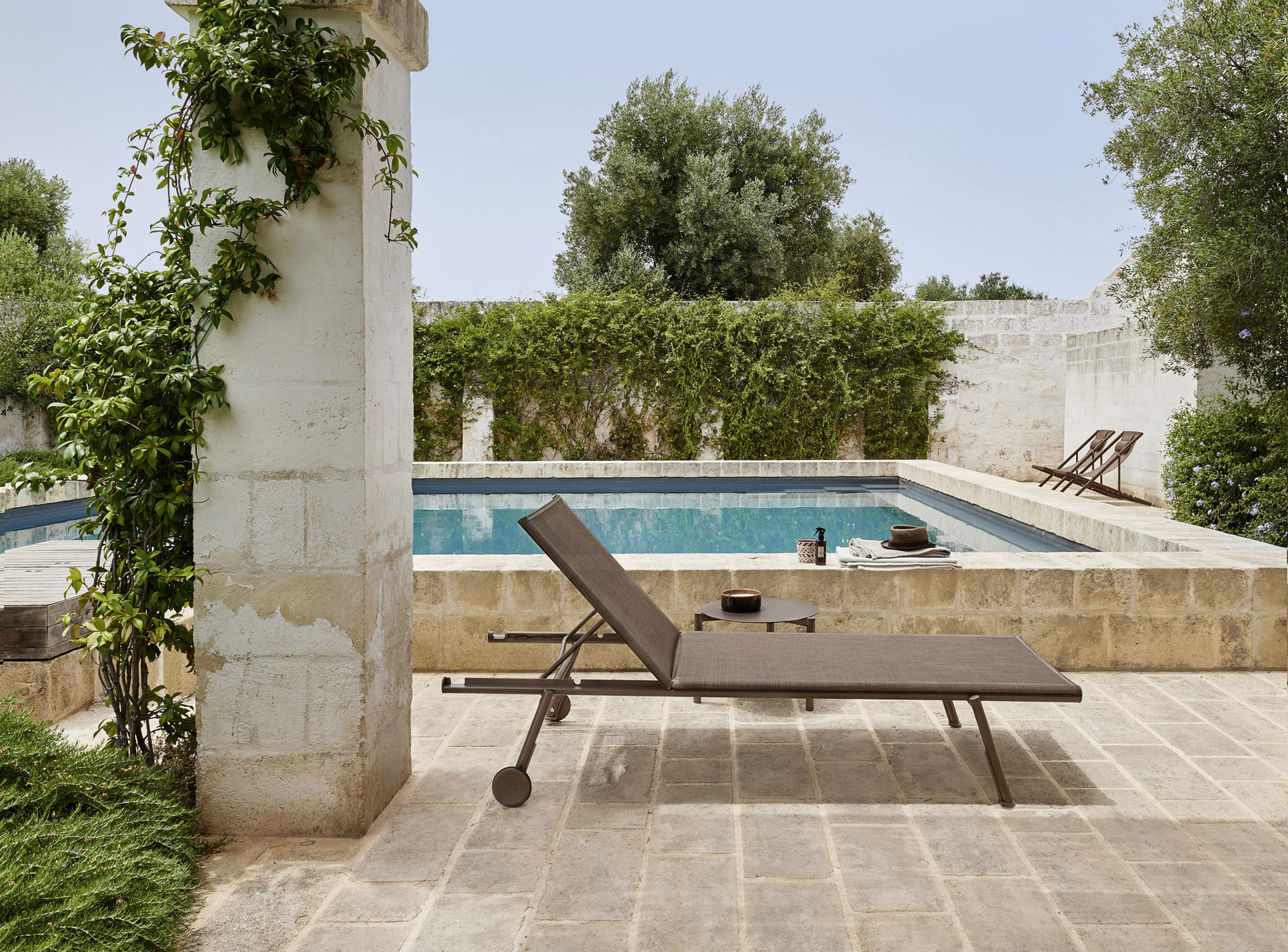 A bronze sling sunlounger and side table on limestone paving beside a rectangular pool in full sun, a vine-covered stone pier at the left edge and dry-stone walling and olive behind.
