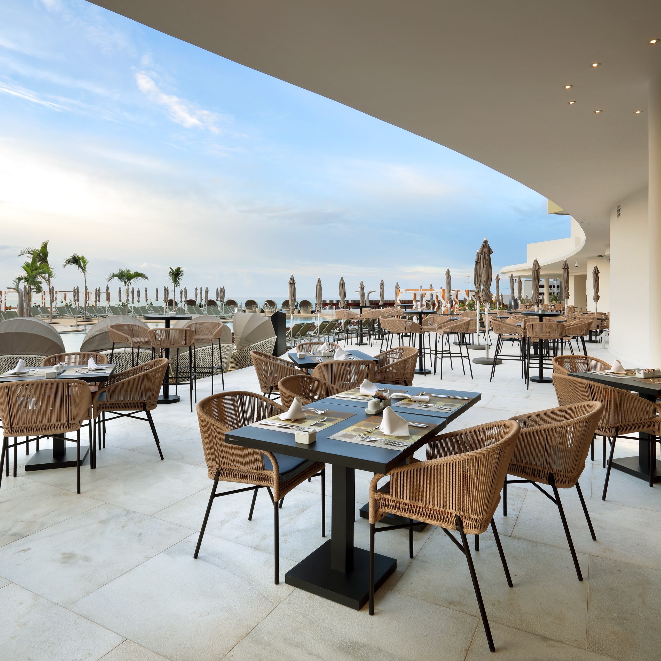 Rope-woven dining chairs at tables set for service on a hotel terrace, with the pool deck and the sea beyond.