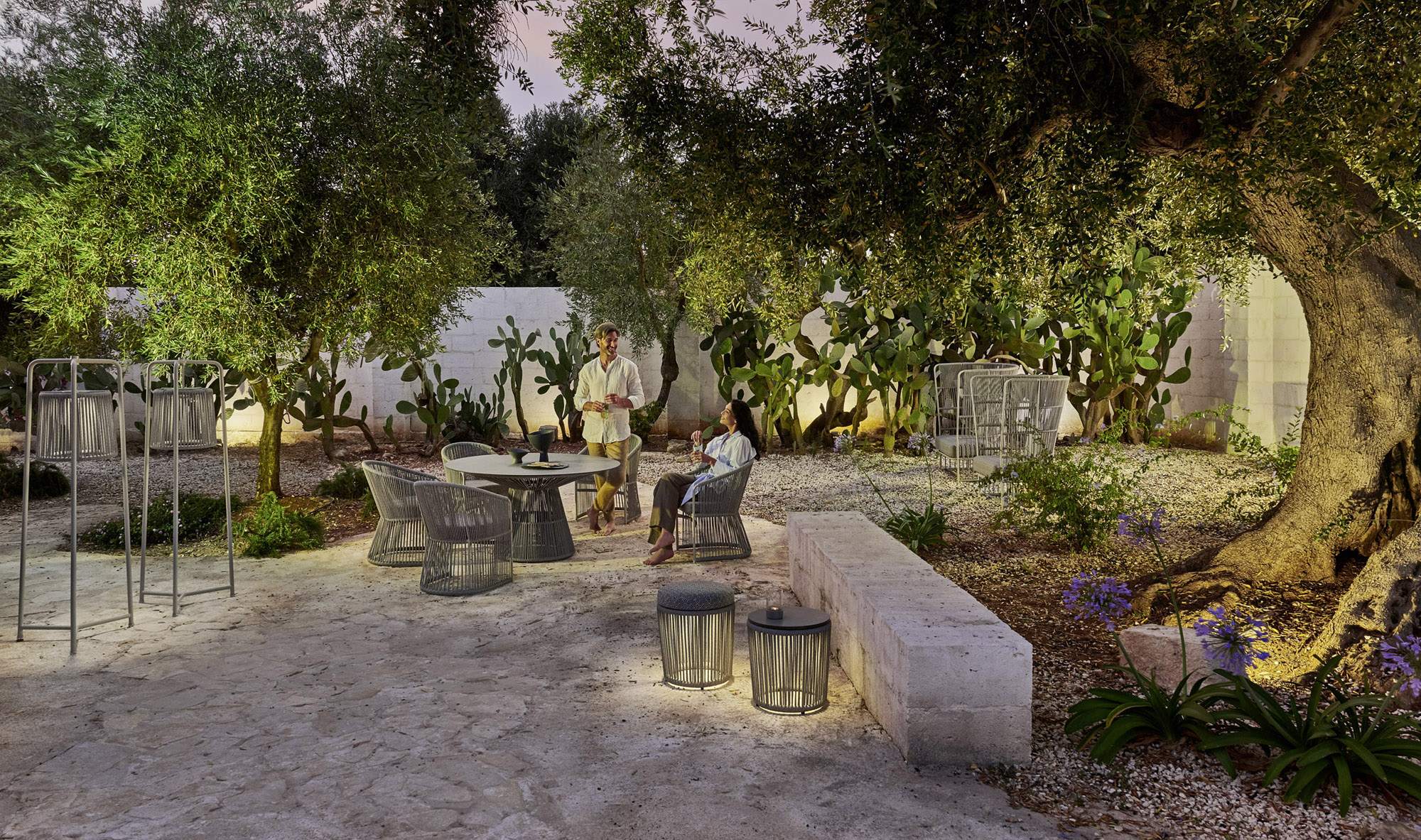 Cord-woven dining armchairs round a table in a gravel courtyard at dusk, two people at the table, olive trees and a bed of prickly pear behind.