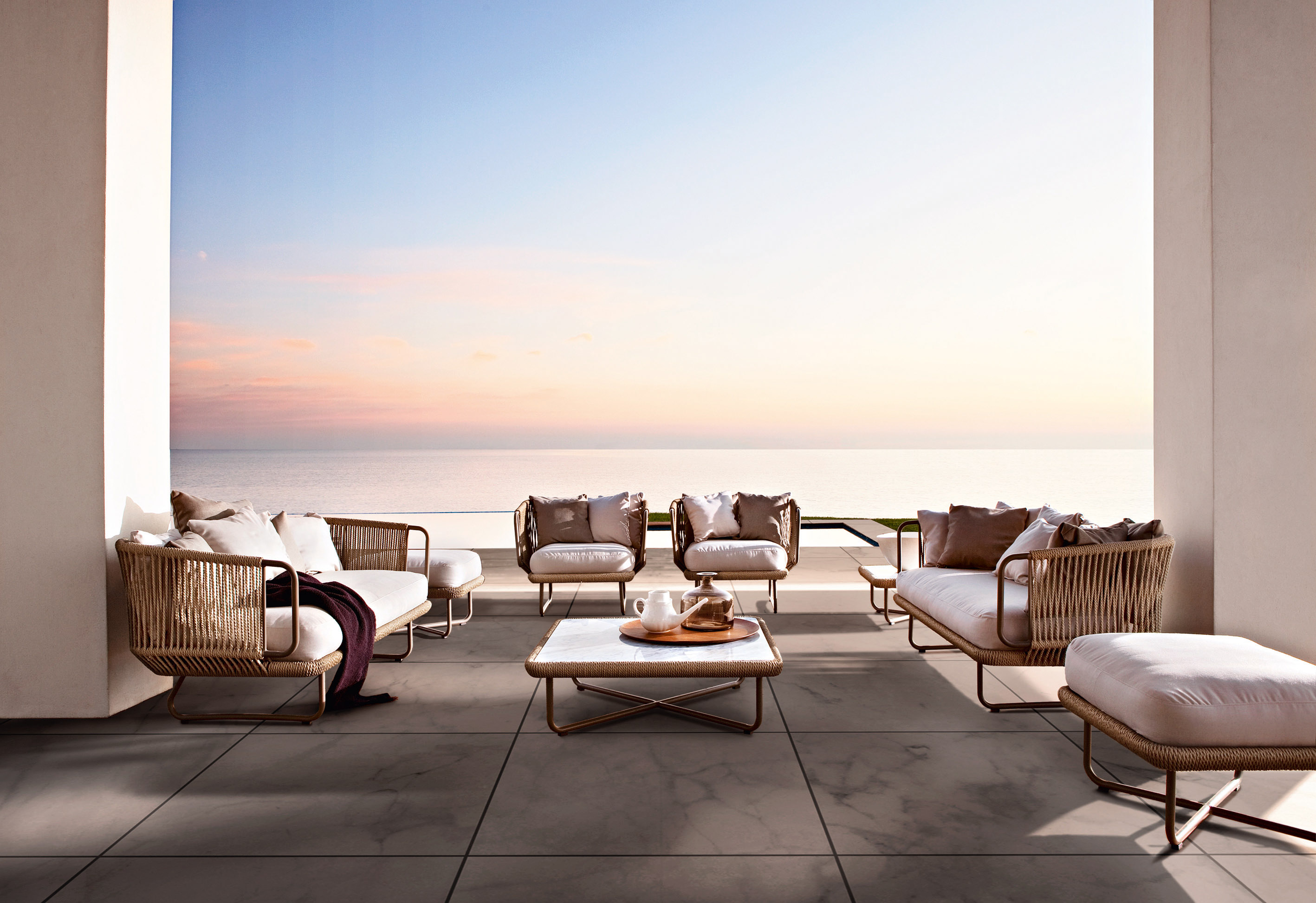 A covered terrace at sunset: woven lounge chairs and a low table facing an open sea horizon.