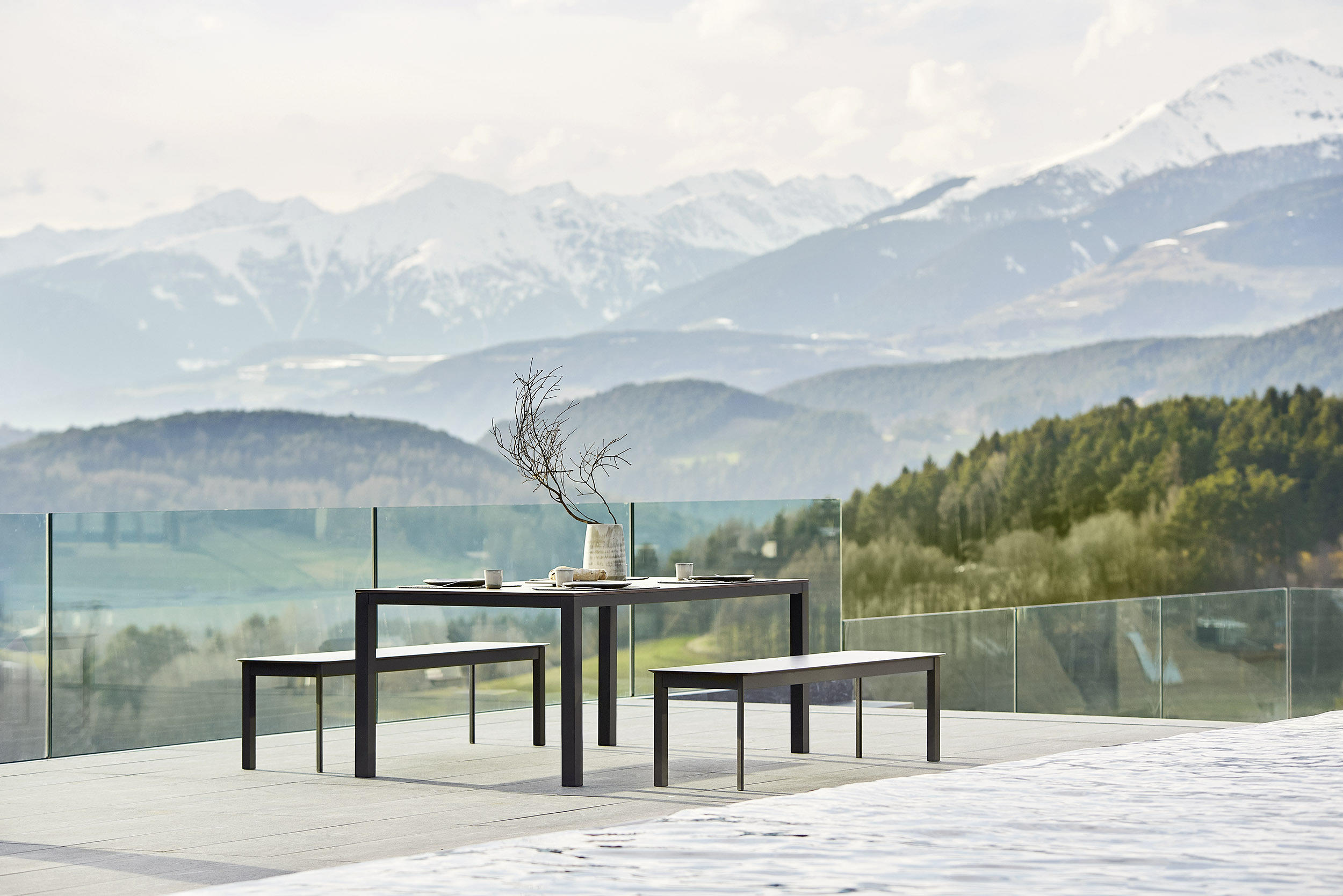 A charcoal-grey dining table and two matching benches with dark HPL tops on a pale stone terrace behind a frameless glass balustrade, a bare branch in a stoneware vase and two places laid on the table, wooded foothills and a snow-capped mountain range beyond in soft alpine light.