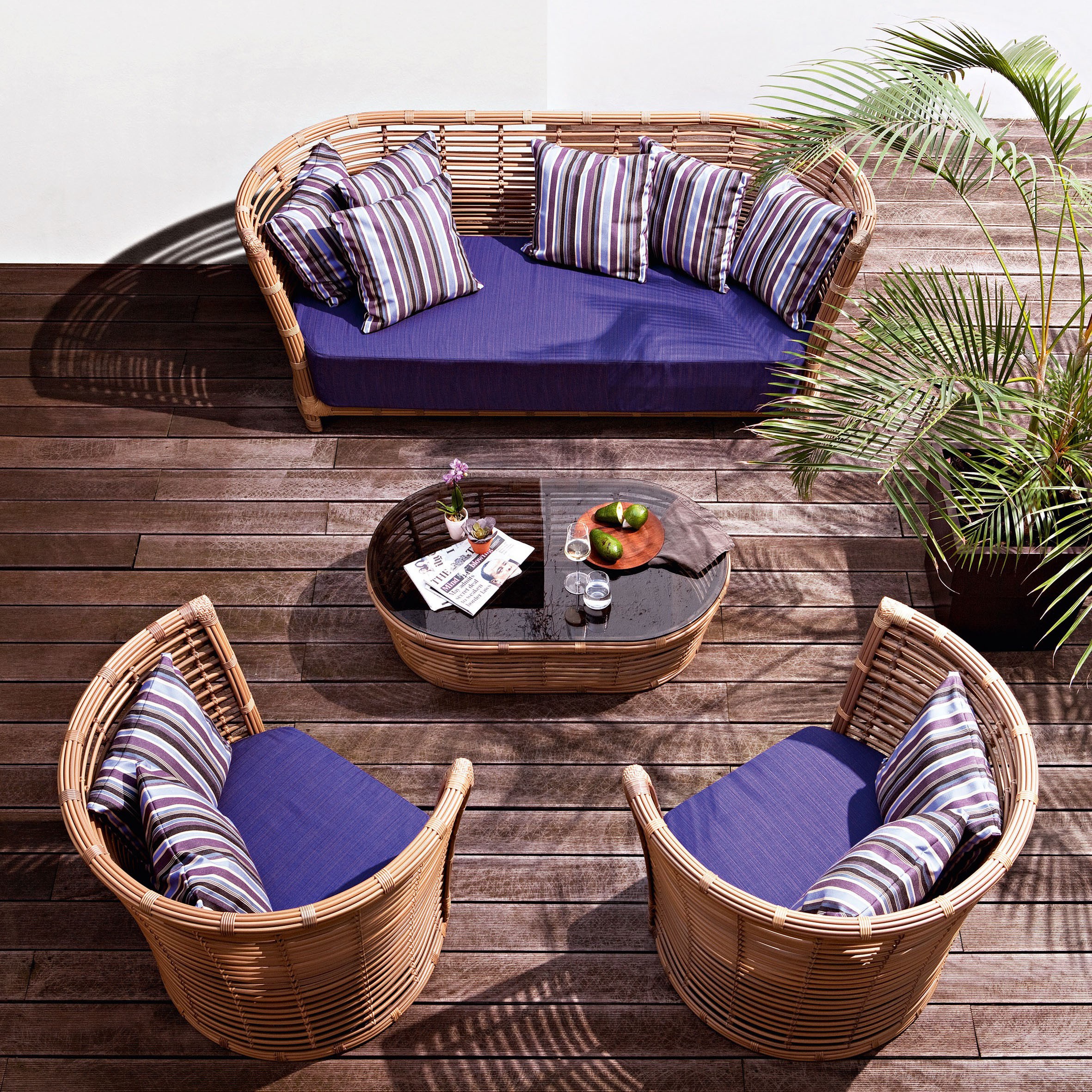 A curved rattan sofa, two barrel chairs and an oval glass-topped table with indigo cushions, on a weathered timber deck.