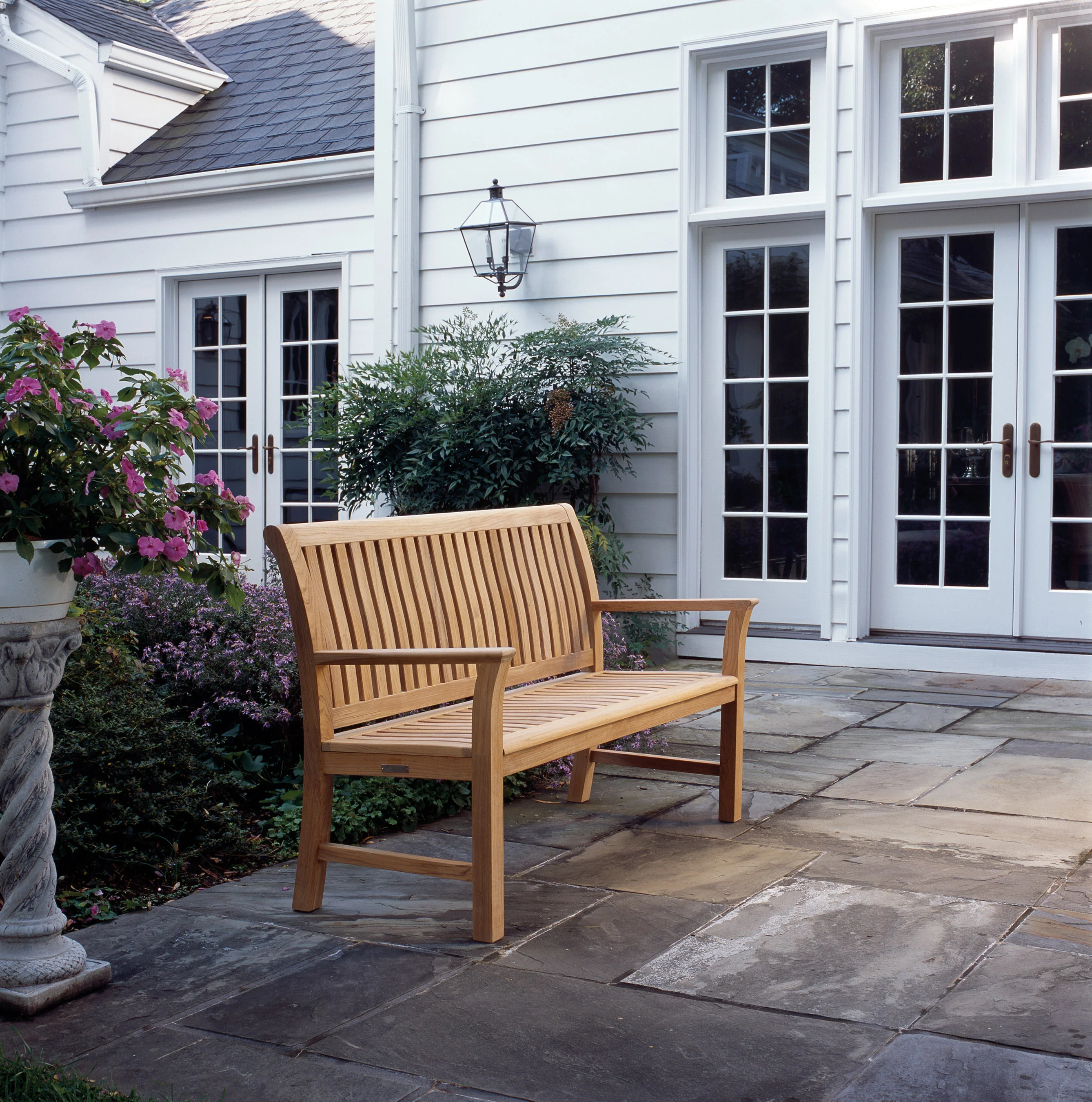 A slatted teak bench on bluestone paving before a white weatherboard house with French doors and a lantern.