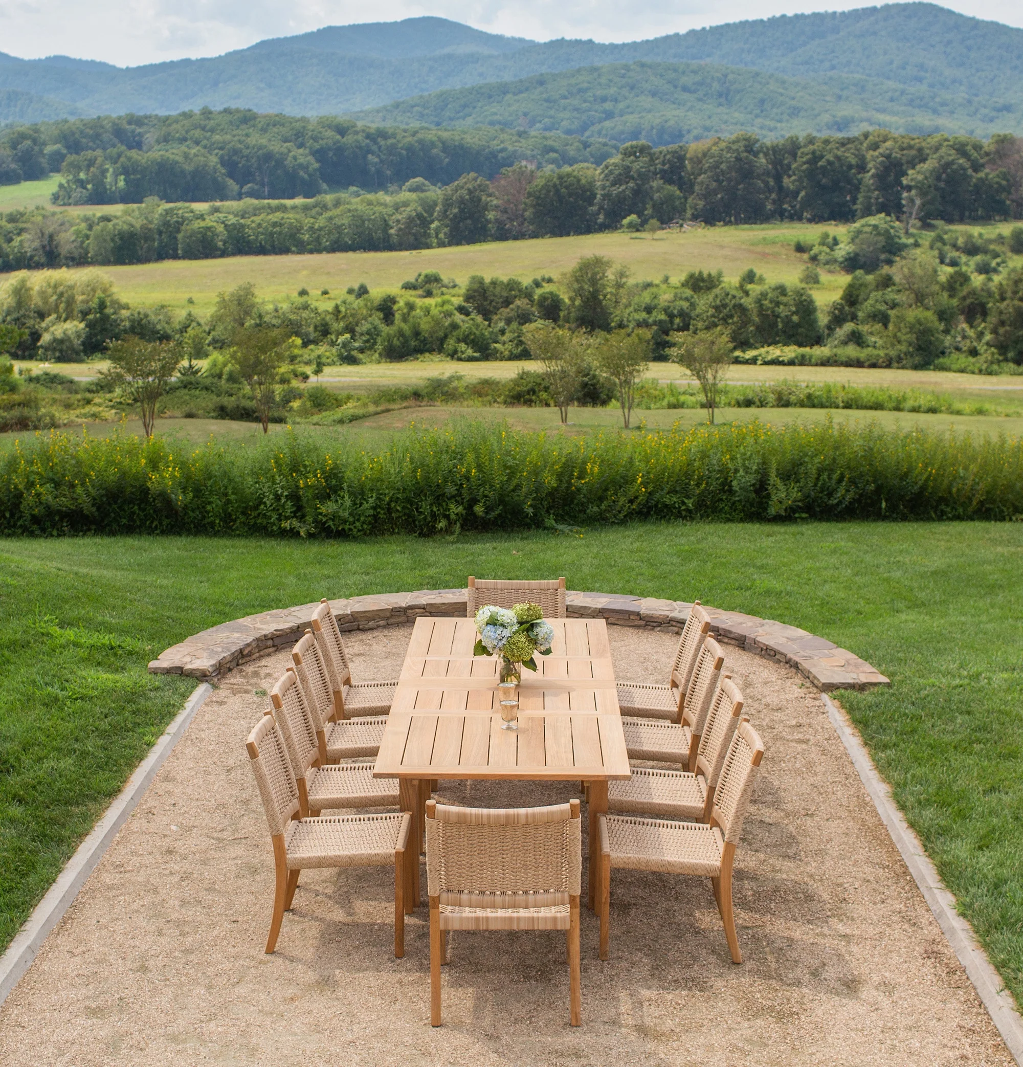 A long teak extension table with a jug of blue and white hydrangeas, rope-woven teak dining chairs around it, on a curved gravel terrace edged with low stone walling above a mown lawn, open pasture falling away to wooded hills and a blue mountain ridge.