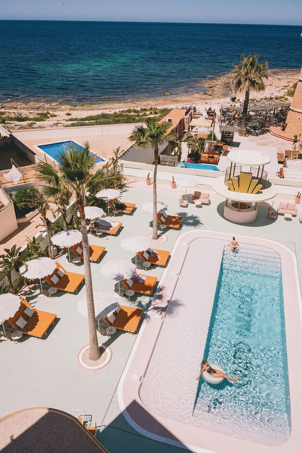 A high view over the hotel from the sea side — beach and open water at the top, then palms, the lap pool and the ranks of orange loungers on the deck below.