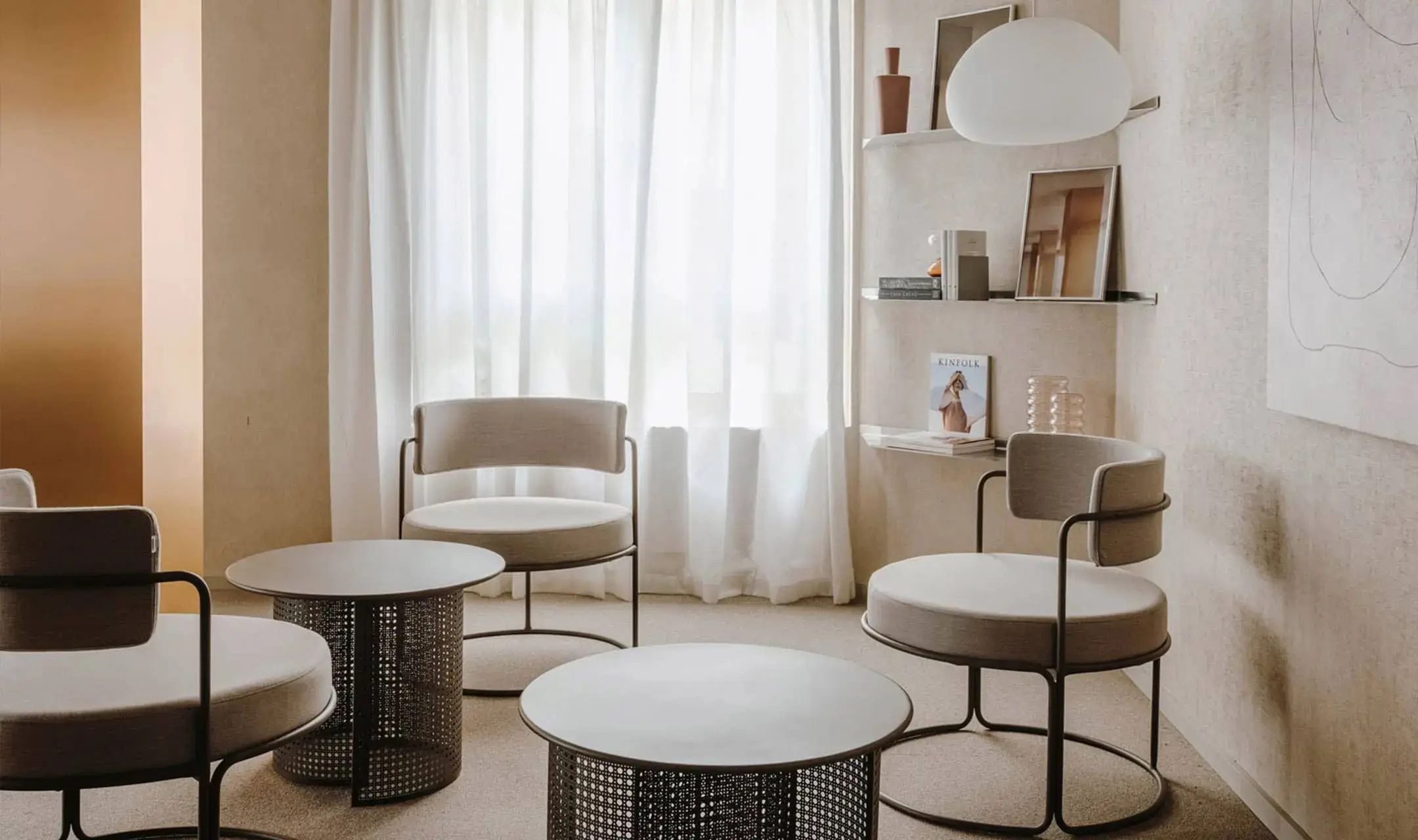 Three low lounge chairs with dark bronze frames and oatmeal upholstered seats and curved backs, grouped around two round-topped coffee tables on dark perforated cane-mesh drum bases, in a pale plastered room with a full-height sheer-curtained window, an ochre wall plane at the left edge and open shelves of books, a vase and framed prints beneath a large white globe pendant.