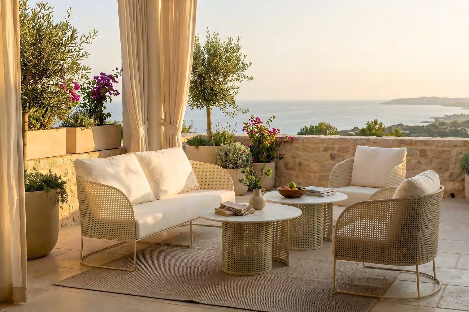 A sand-toned perforated-steel two-seat sofa and two lounge chairs with ivory cushions around a pair of round drum-based tables on a stone terrace, cream curtains and potted olive trees at the left edge, bougainvillea against a low stone wall beyond, a sea horizon in warm golden-hour light.