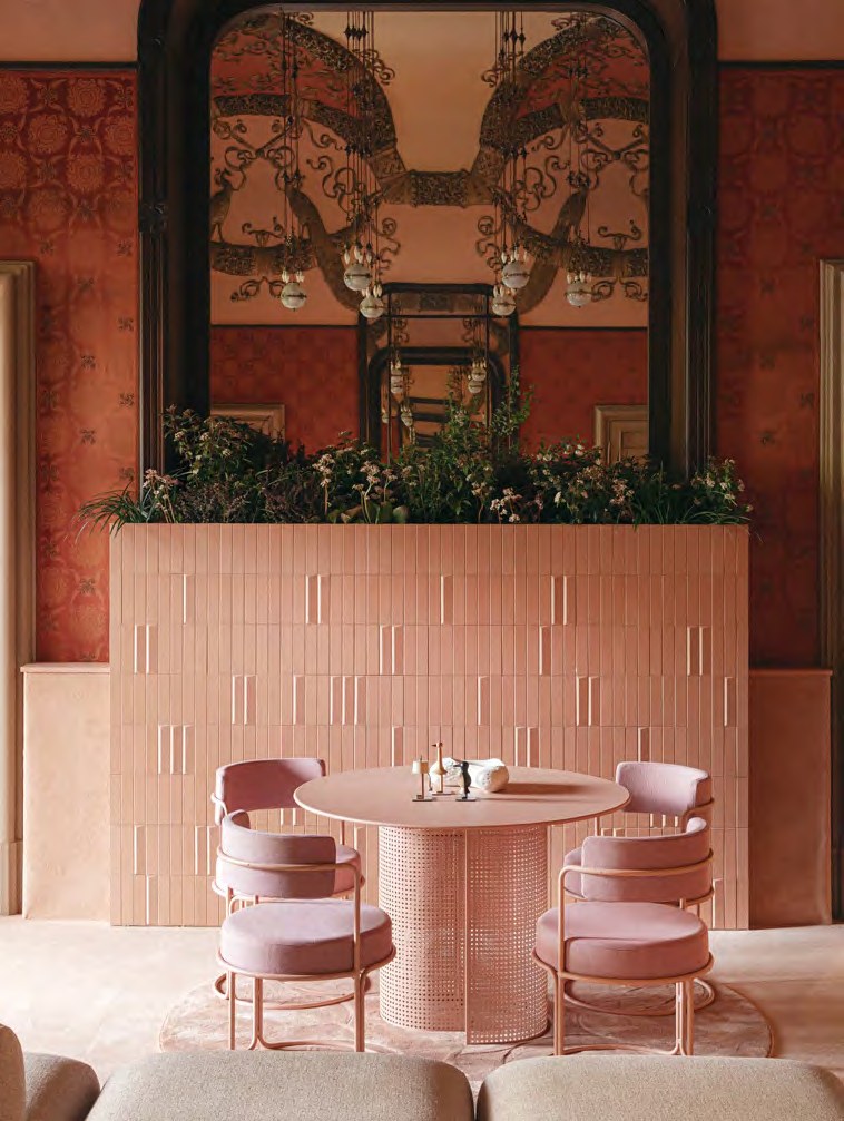 A round pastel-pink dining table on a perforated cylindrical base with four matching pink armchairs around it, set against a pale pink glazed-tile bar counter topped with trailing planting, a tall arched mirror behind reflecting an ornate belle-epoque ceiling and chandeliers, deep terracotta damask walls either side.