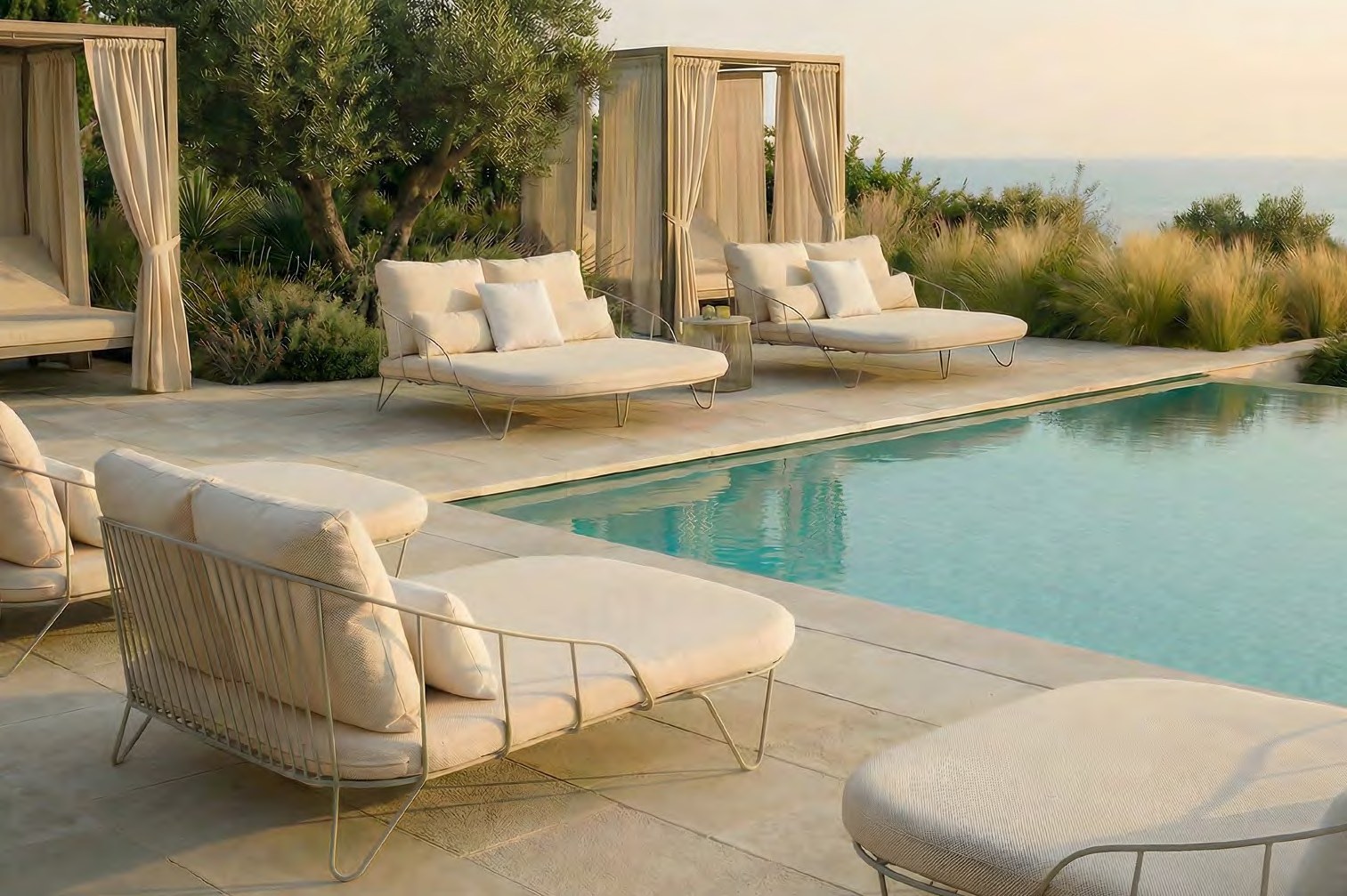 Pale wire-framed OLIVO daybeds with ivory cushions grouped beside a rectangular infinity pool, curtained cabana structures and an olive tree behind them, ornamental grasses and a sea horizon beyond, in warm late-day light.